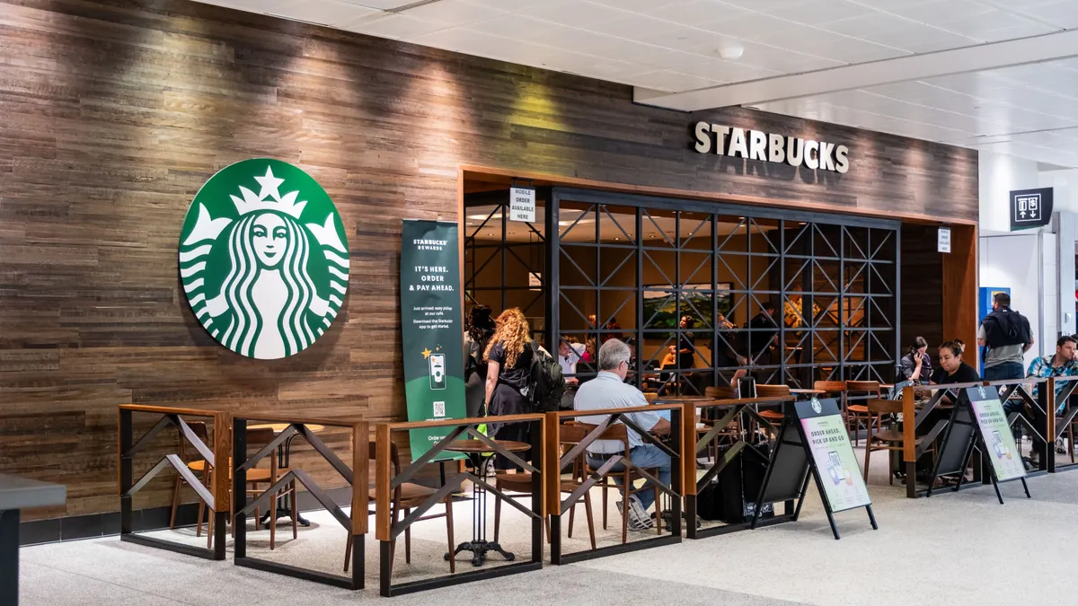 An image of a small Starbucks storefront with brown paneling and green siren logo inside an airport. Several people are sitting in chairs with luggage.