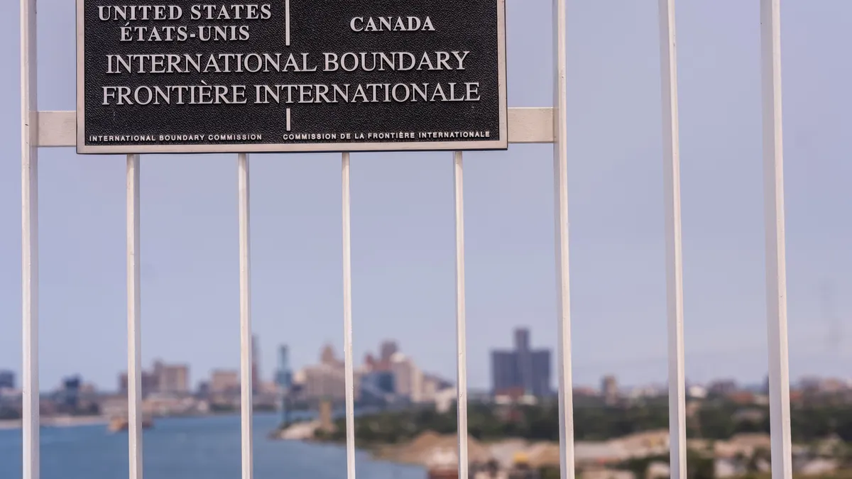 Metal sign that says "United States Etats-Unis Canada International Boundary Frontiere Internationale" on metal bars with body of water and city skyline behind it.