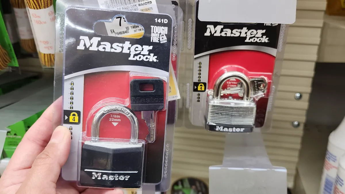 A person holding a black Master Lock in its clear plastic packaging, with another Master Lock package in silver behind it.