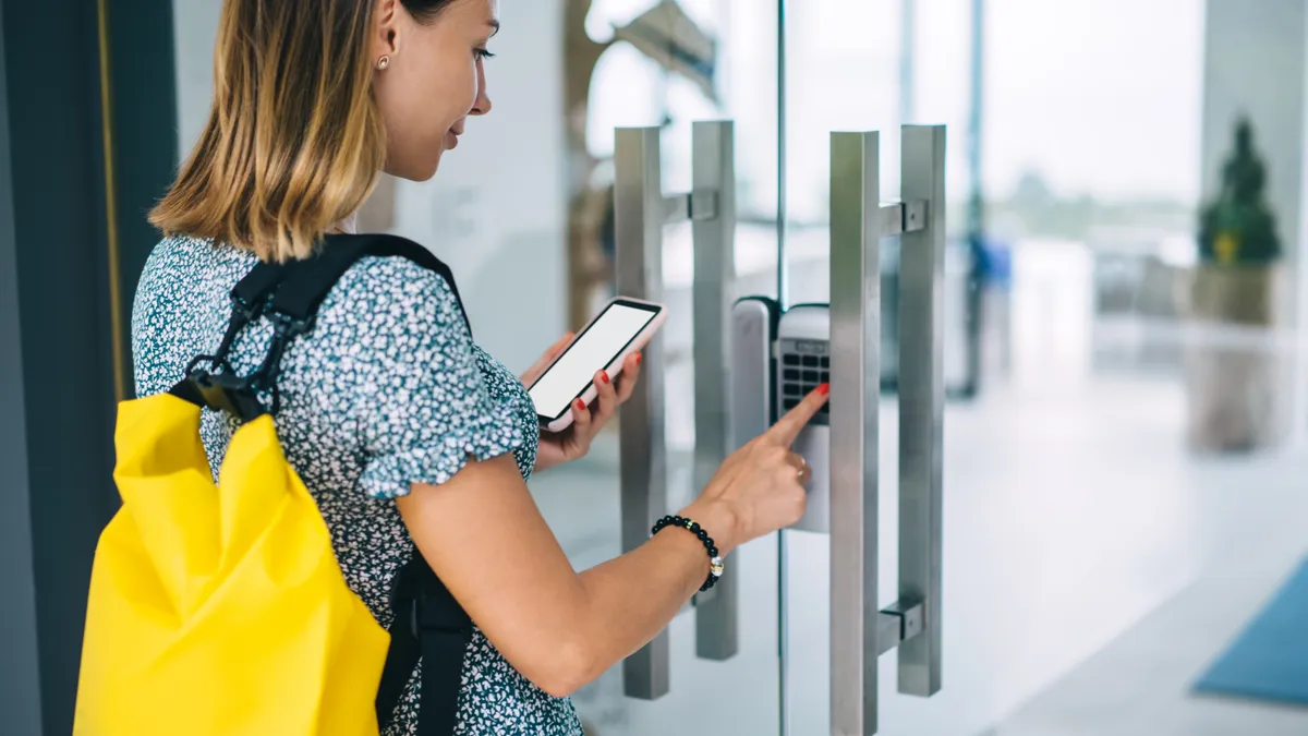 A female with a backpack standing near a doorway access control panel and using a mobile phone to enter a security code.