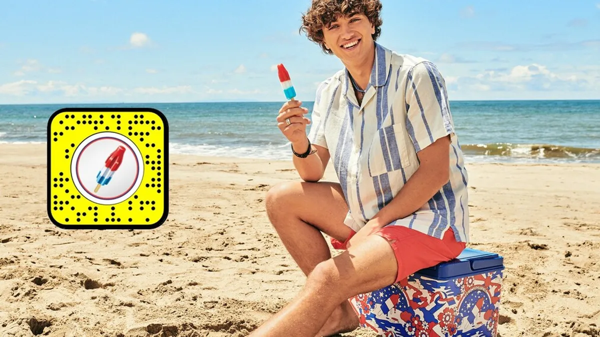 Actor Gavin Casalengo sits on a beach holding a Bomb Pop in one hand.