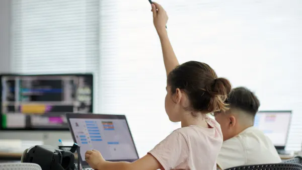 A young student raises their hand in a classroom while sitting at a desk and using a laptop.
