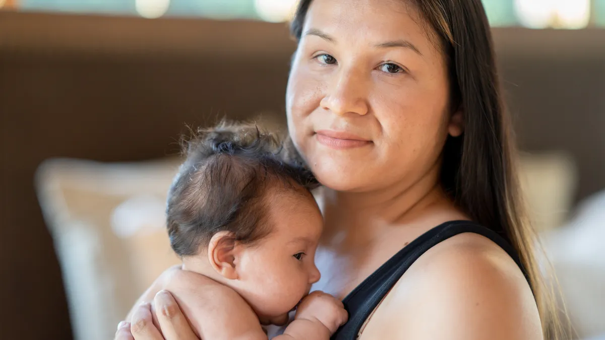 A new mother of Native American descent holds her newborn baby to her chest. The mother is wearing a black tank top and looking at the camera with a gentle smile.