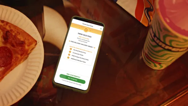 A photo of a smartphone on a table with pizza on a plate next to it on the left and a cup of soda on the right. The mobile app says "7Now Gold Pass. First 14 days free. Renews automatically. Cancel anytime. Free delivery on every order and extra perks for orders over $10. No small basket fees, 2x rewards points, FREE product on every order. Additional member perks"