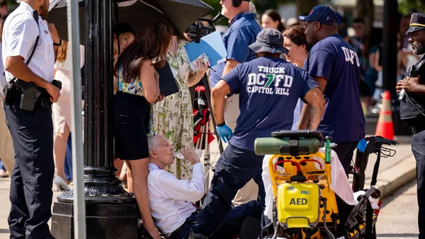 People crowd around a person sitting on the ground drinking water out of a plastic bottle. One person holds an umbrella over the person on the ground.