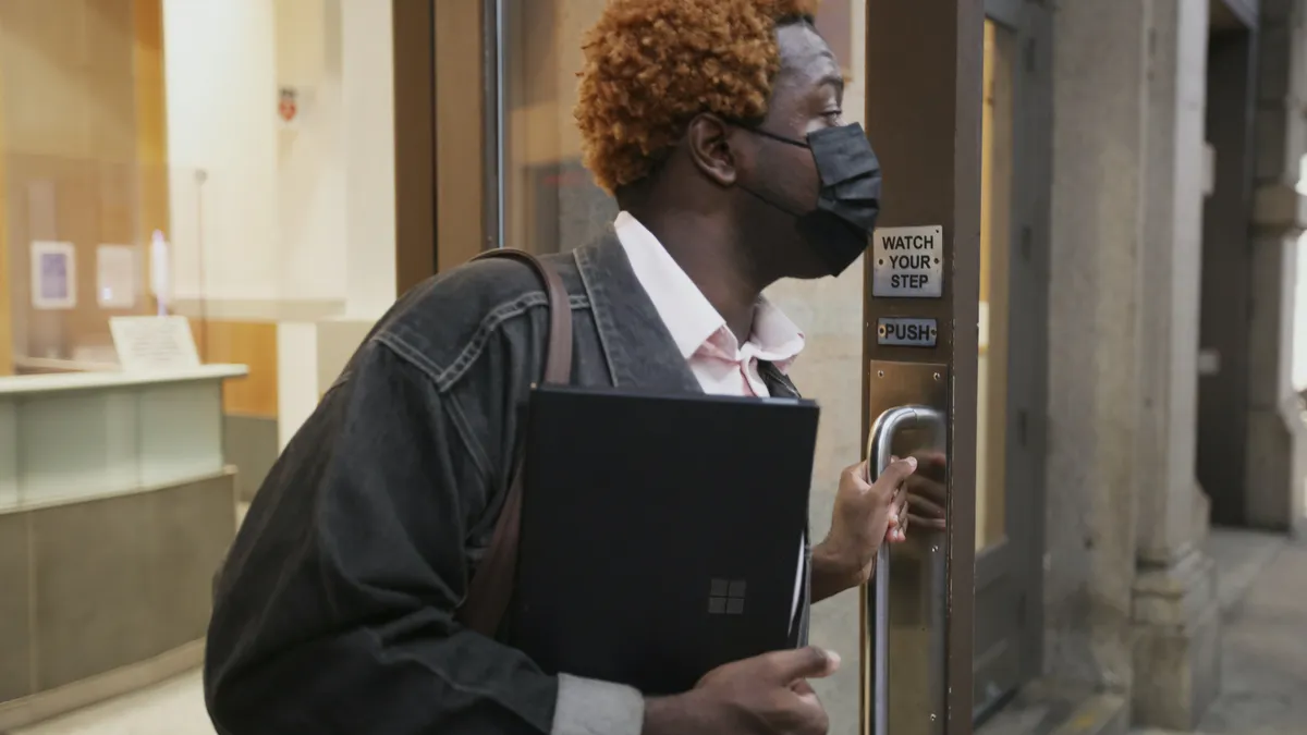 A Black person exits an office; holds a laptop