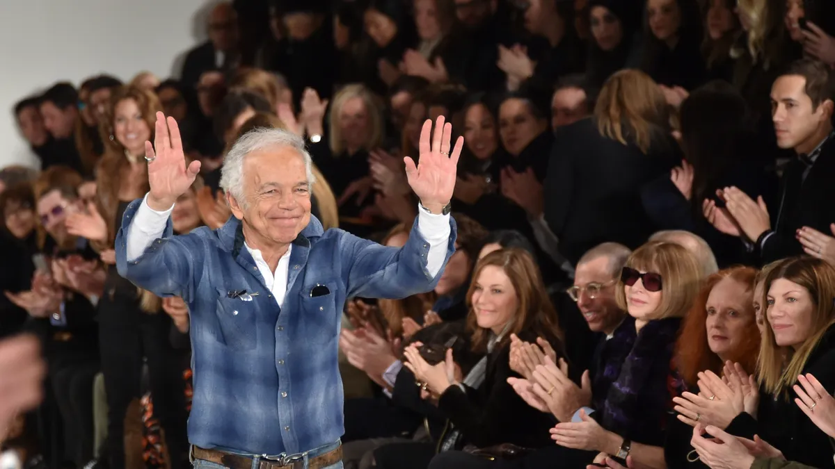 Ralph Lauren wears jeans and a blue plaid shirt over a long sleeved white shirt and stands on the end of a fashion runway showing his palms to the applauding crowd and smiling