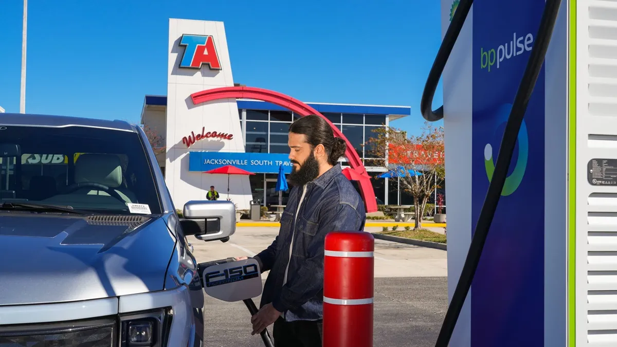 A photo of a person plugging an EV charging plug into a vehicle. There is a building in the background with signs that say "TA," "Welcome" and "Jacksonville South travel center." The EV charger has wording that reads "BP Pulse."