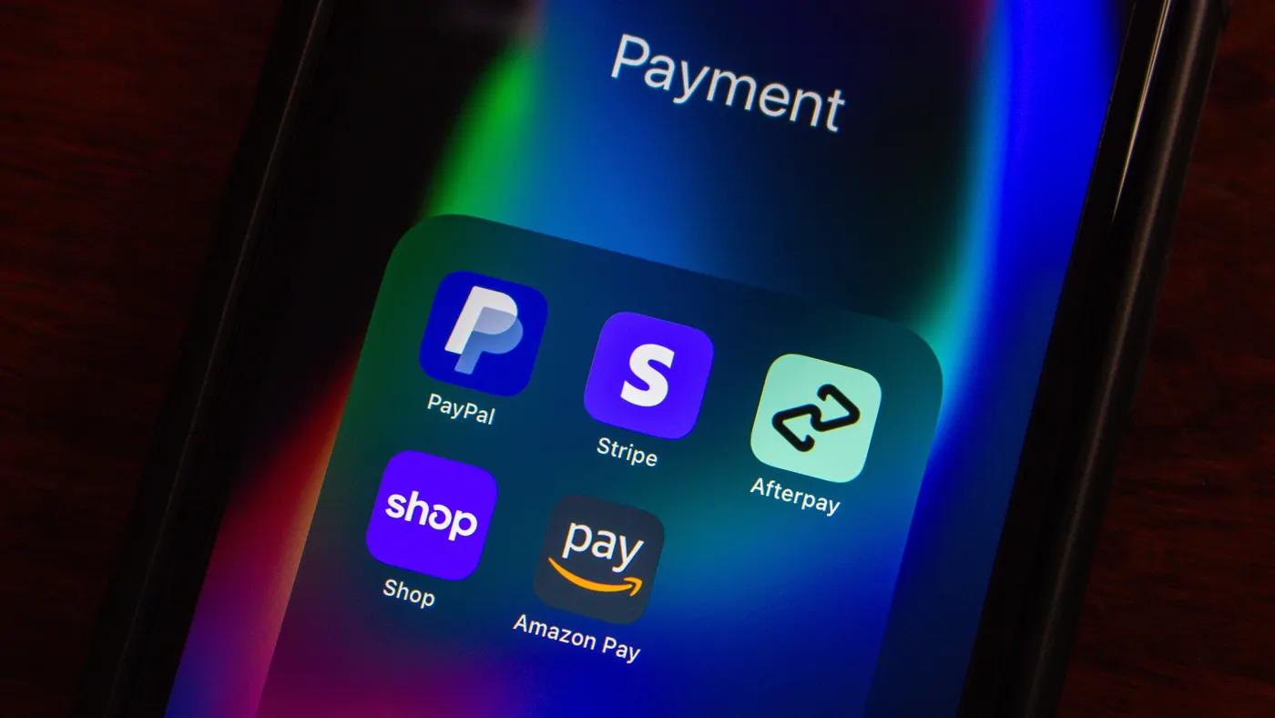 A smartphone screen displaying a folder labeled “Payment” with apps including PayPal, Stripe, Afterpay, Shop, and Amazon Pay, shown in a dark setting with colorful screen reflections.
