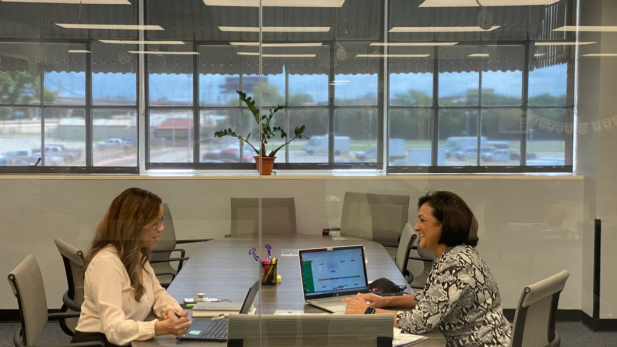 Two women sit on opposite sides of a conference table facing each other.