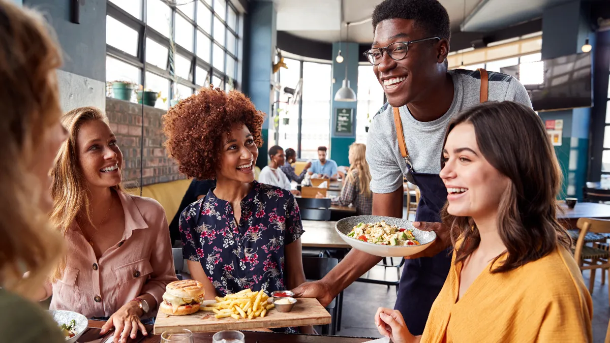 A male server serves four customers sitting at a table with a bowl of pasta and a hamburger