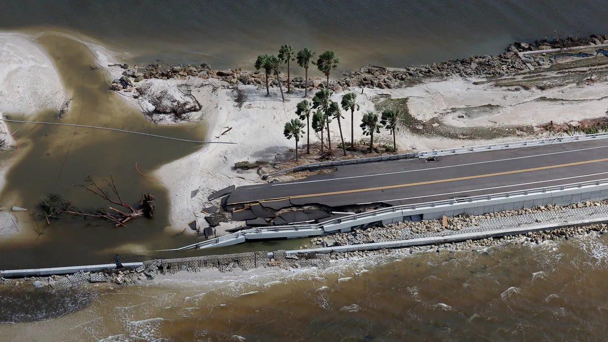 An aerial view shows ocean overtaking a road, which has cracked asphalt and crumpled railings.