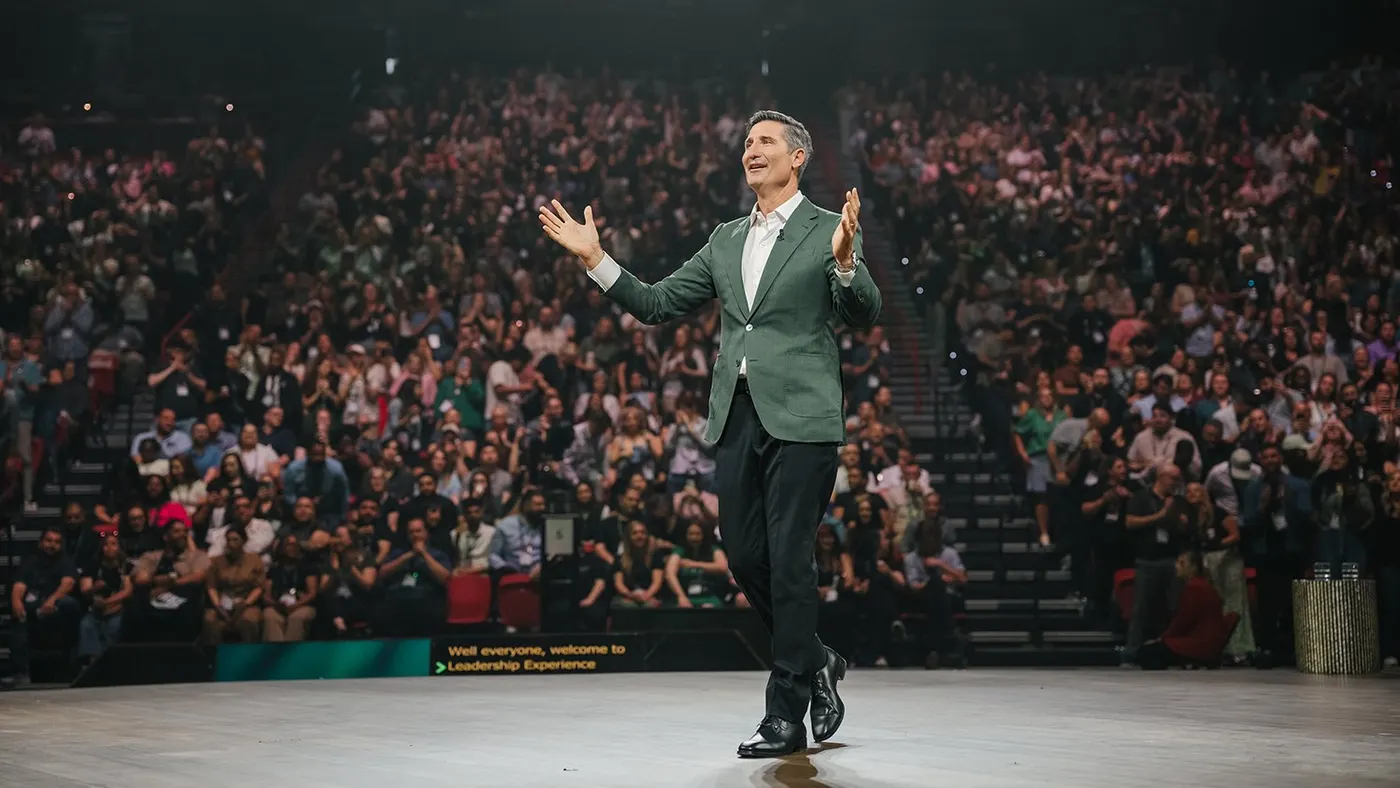 A person in a suit stands on stage surrounded by a large crowd in the stands.