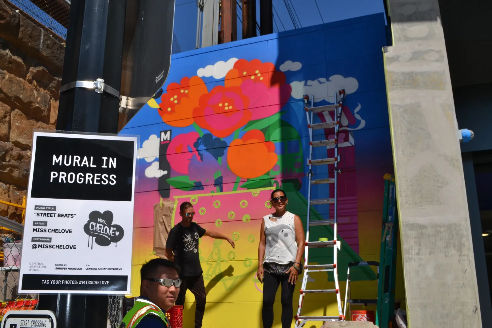 Two people stand in front of a colorful mural. A sign reads "Mural in Progress."