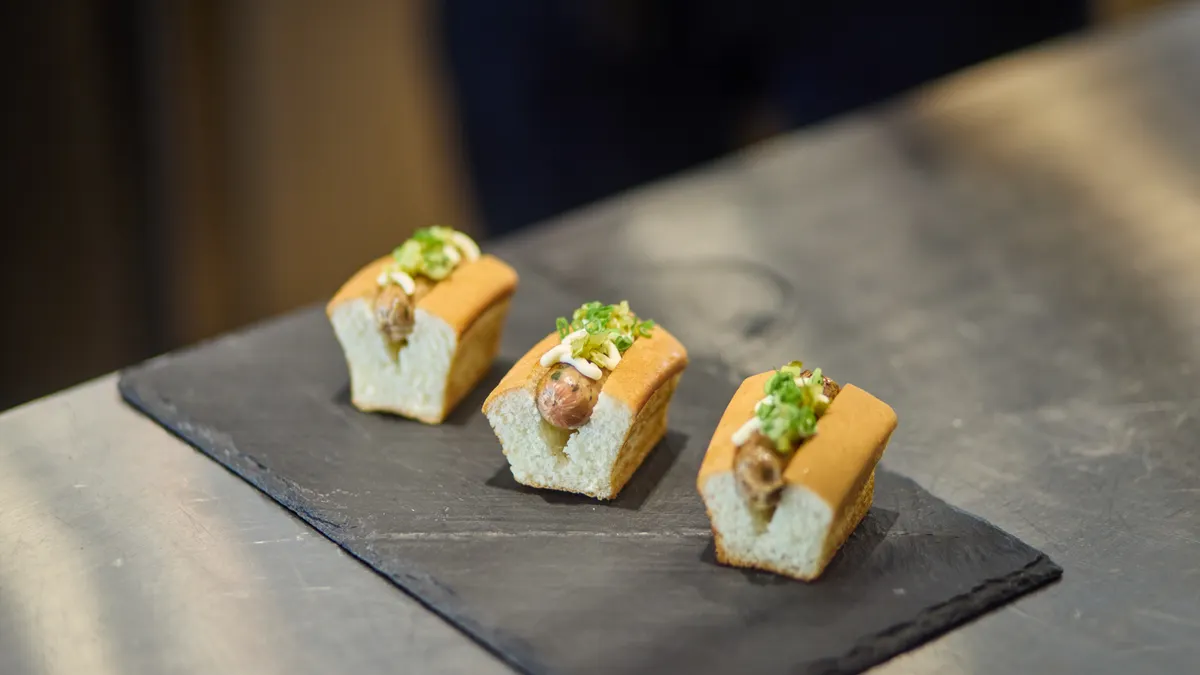 Three small sausages made of cultivated meat sit in V-shaped buns with drizzles of white sauce and sprouts on top. They are sitting on a piece of slate on top of a metal table.