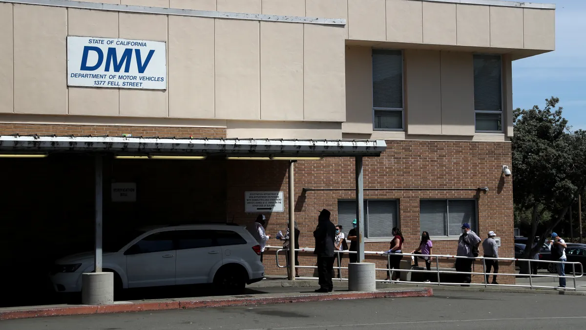 People line up to enter a low building with a sign identifying it as a California Department of Motor Vehicles office. A white car is parked under an awning in front of the building.