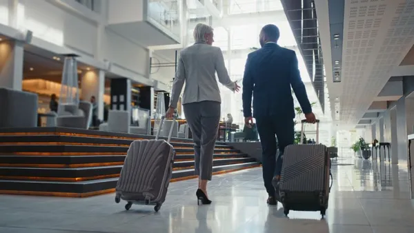 Business travelers walk through hotel lobby carrying luggage.