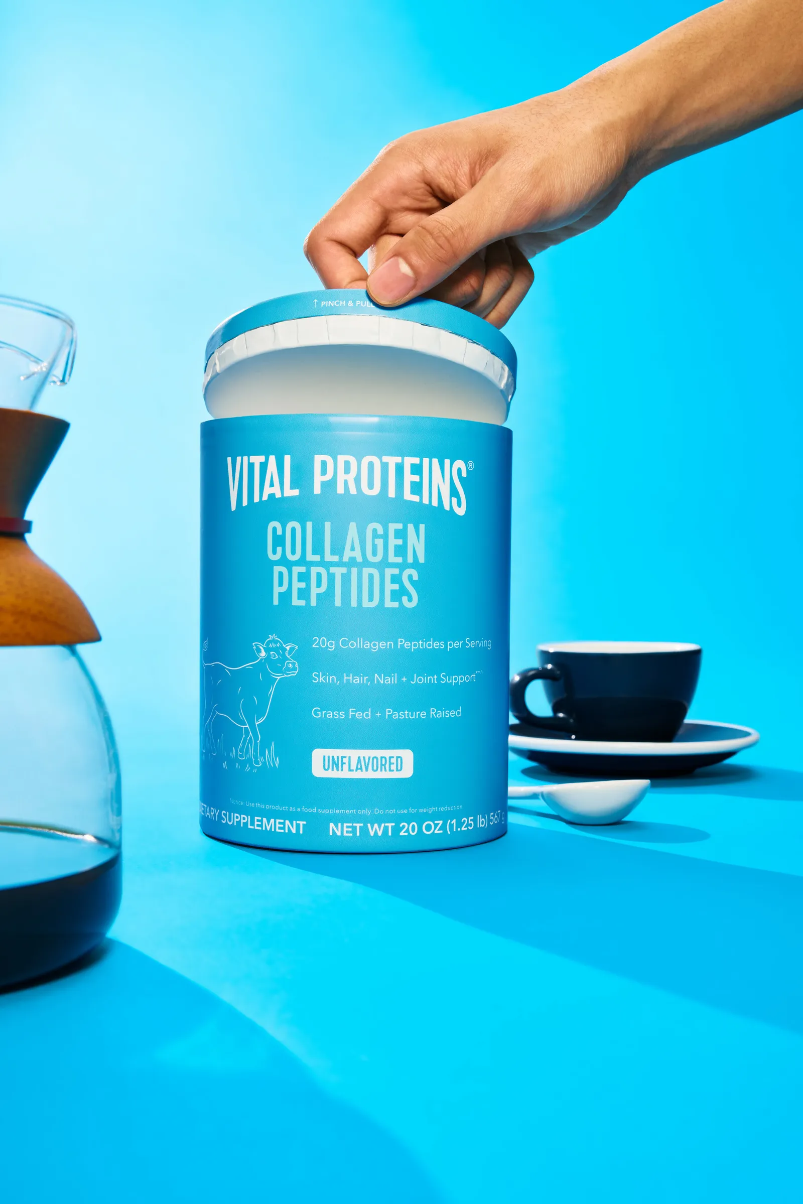 A person&#x27;s hand opens the lid of a blue and white paper-based canister for Vital Proteins collagen peptides.