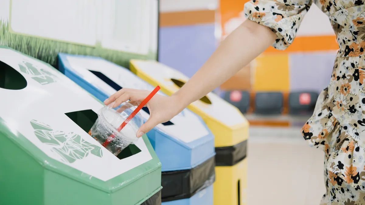 A person drops a clear plastic cup into a public recycling bin.