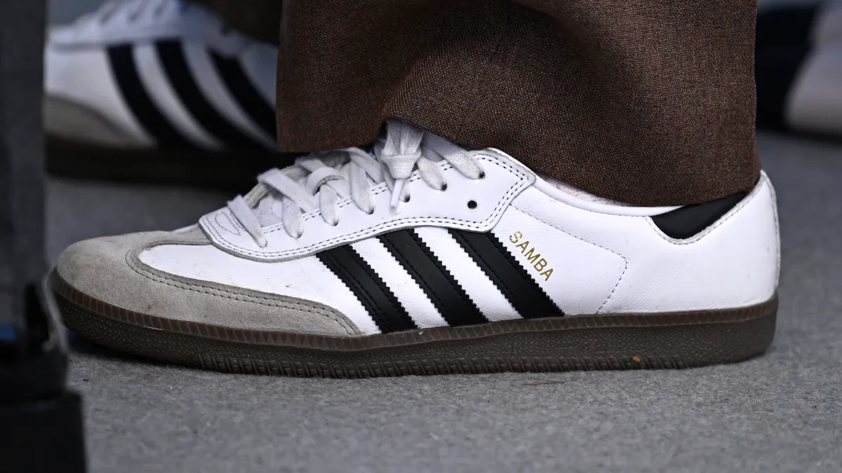 A closeup of an actor's shoes at a red carpet premerie. They are white Adidas Sambas with black stripes and a grey toe.