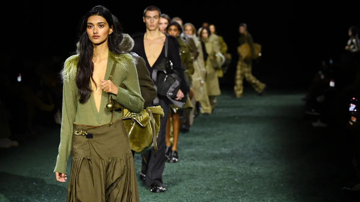 Models in neutral toned outfits walk down a darkened runway.