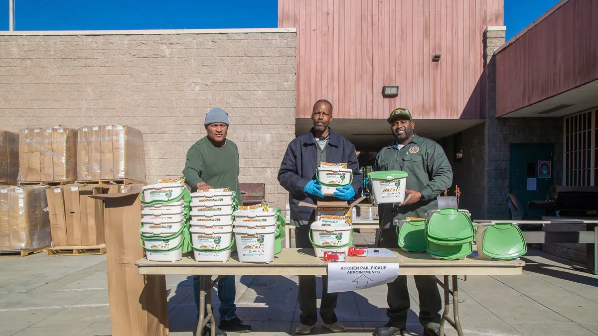 Men in workwear hold plastic kitchen pails for composting behind a folding table