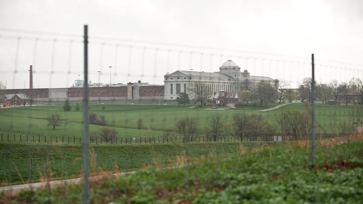 Distant view of a grey building with a flag brick facility attached and grassy hills in the foreground, crossed by wire fencing.