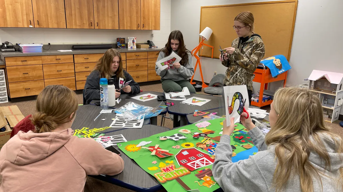 A group of girl high school students gathers around a table to cut out designs for an elementary school makerspace.