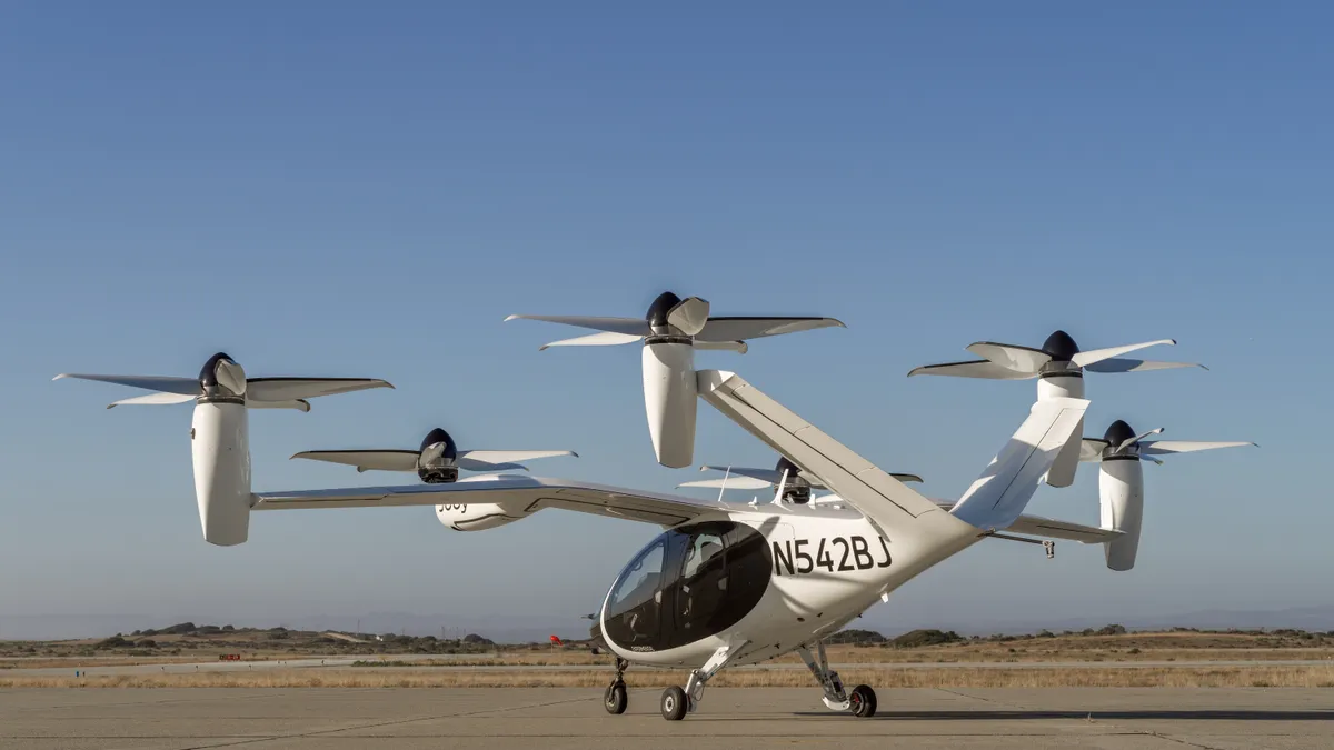 A helicopter-shaped futuristic white aircraft with six upright propellers sits on a concrete pad.