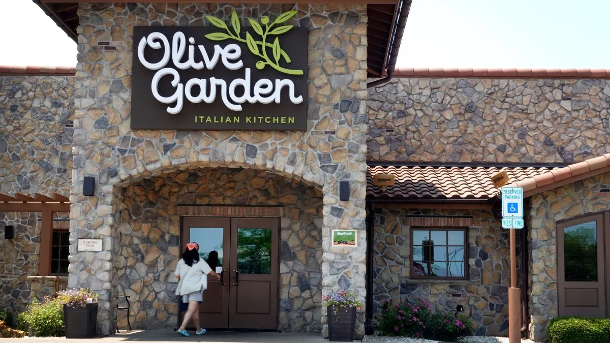An iamge of a stoneworked building with a white logo that says Olive Garden. A woman wearing a white shirt and red bandana is at the door about to enter the restaurant.