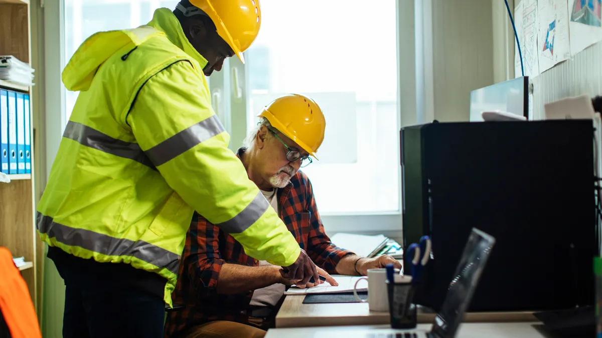 Two workers in hardhats look at blueprints in an office.