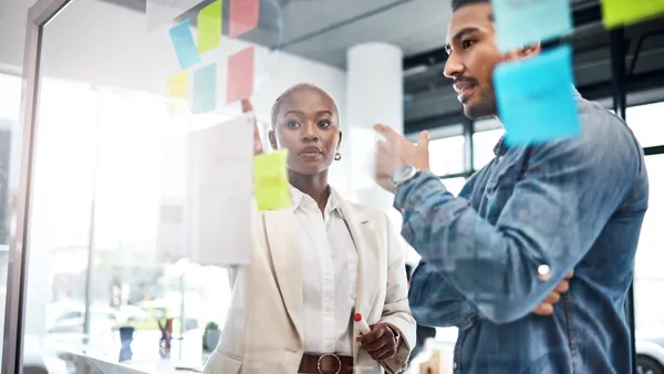 Two people in business attire stand front of a glass wall in an office with sticky notes on it, having a conversation.