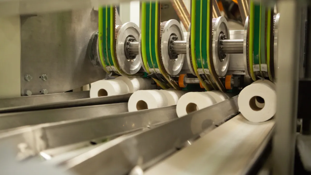 Rolls of toilet paper proceed along a manufacturing line.