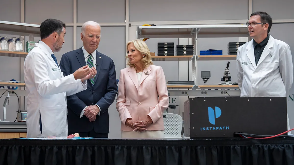 Two people wearing suits stand in between two people talking wearing white coats in a laboratory setting.