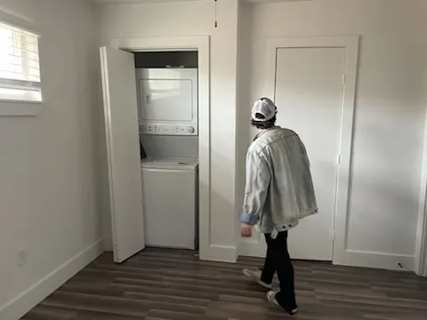 A person walks around an empty room with a window. A washing and drying machine are seen in an open closet.