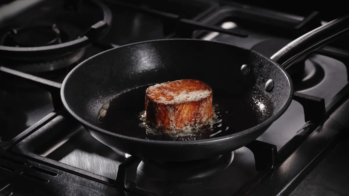 A black frying pan on a black gas range with a sizzling Juicy Marbles Thick Cut Filet cooking inside.