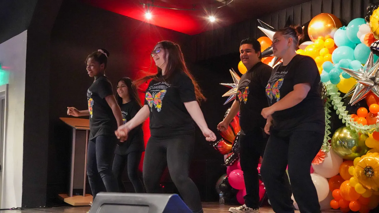 Five members of a high school dance team are shown performing on a brightly decorated stage during a fundraiser for the school's center for the arts.