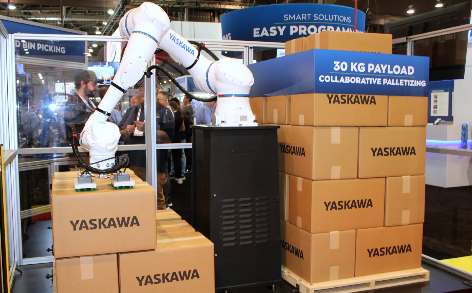 A white robot arm picks up cardboard boxes at a booth display at a conference.