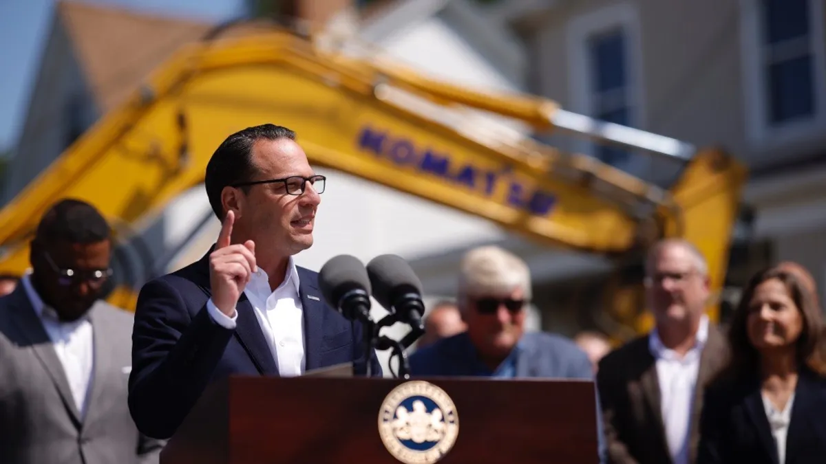 A man speaks at a podium with out of focus construction machinery behind him.