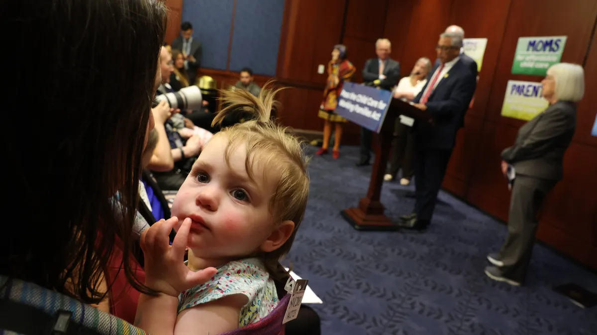 A mom holds a baby while lawmakers speak at a podium in the background.