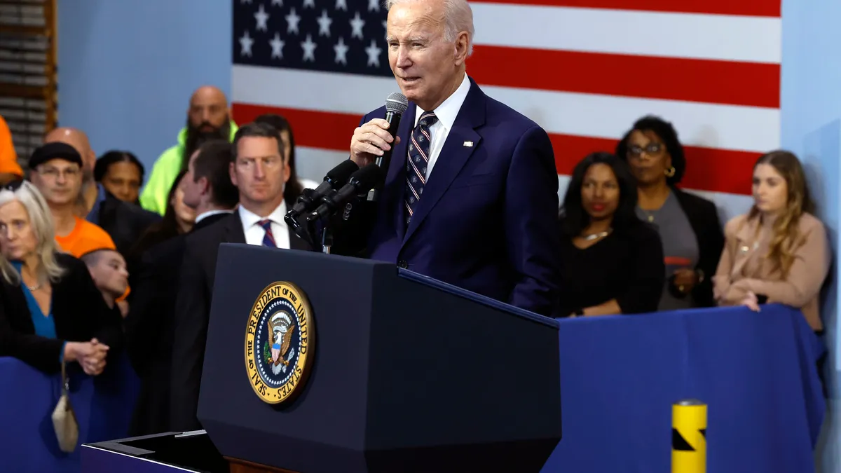 U.S. President Joe Biden in a dark blue suit with a microphone standing behind a podium, with an American flag behind him.