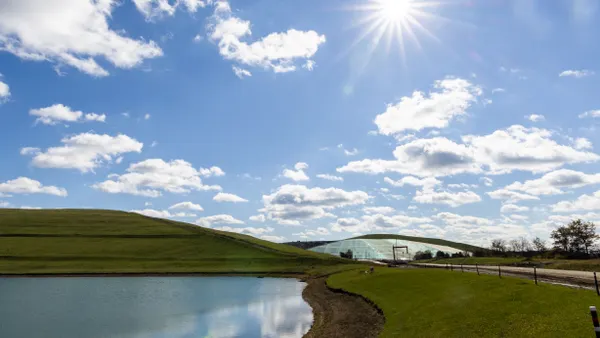 A retention pond in the foreground reflects a partly cloudy sky. Behind it are a grassy hill and a further hill covered in a tarp-like material.