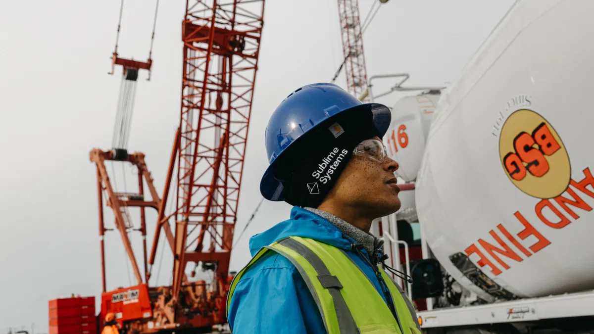 A worker in a blue hard hat and reflective vest stands near a concrete truck, with other construction equipment in the background.