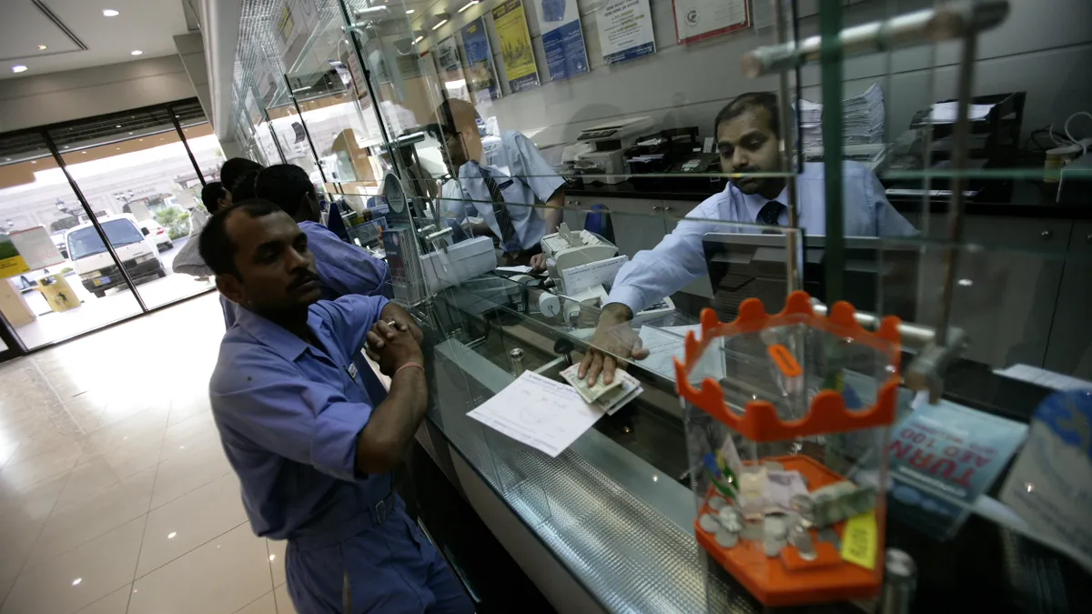 Worker sits at counter in establishment with money and papers in front of him interacting with an employee.