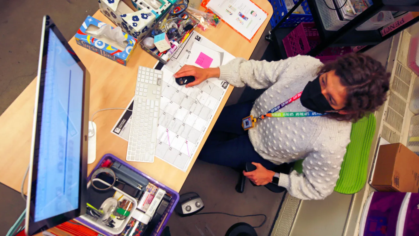 Person is seen from above wearing a face mask and sitting at a desk with a keyboard and monitor. The desk also has a big calendar, a tissue box and other items.