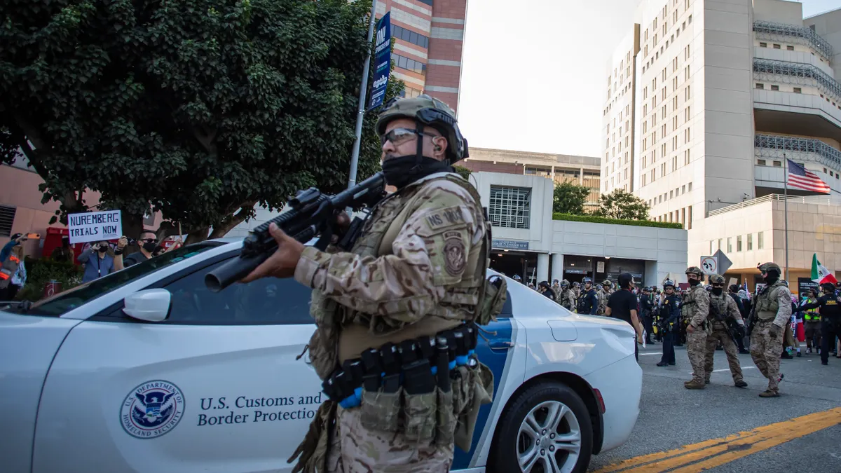 A person in military fatigues stands aiming a gun in front of a car that says "U.S. Customs and Border Protection" with crowd and military officers in the background.