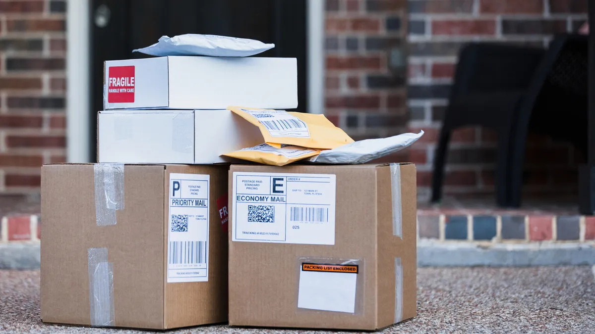 Mailed packaging pouches rest atop cardboard box packages on the front porch in front of a home