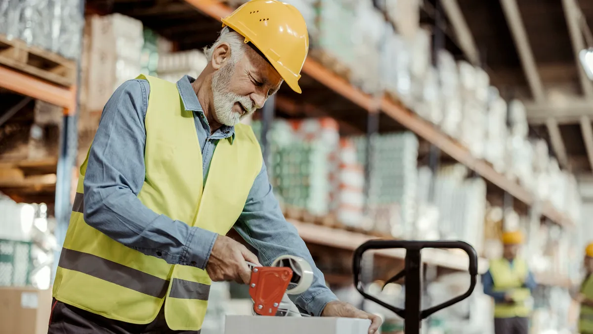 Senior male worker packing cardboard box with tape gun dispenser in warehouse