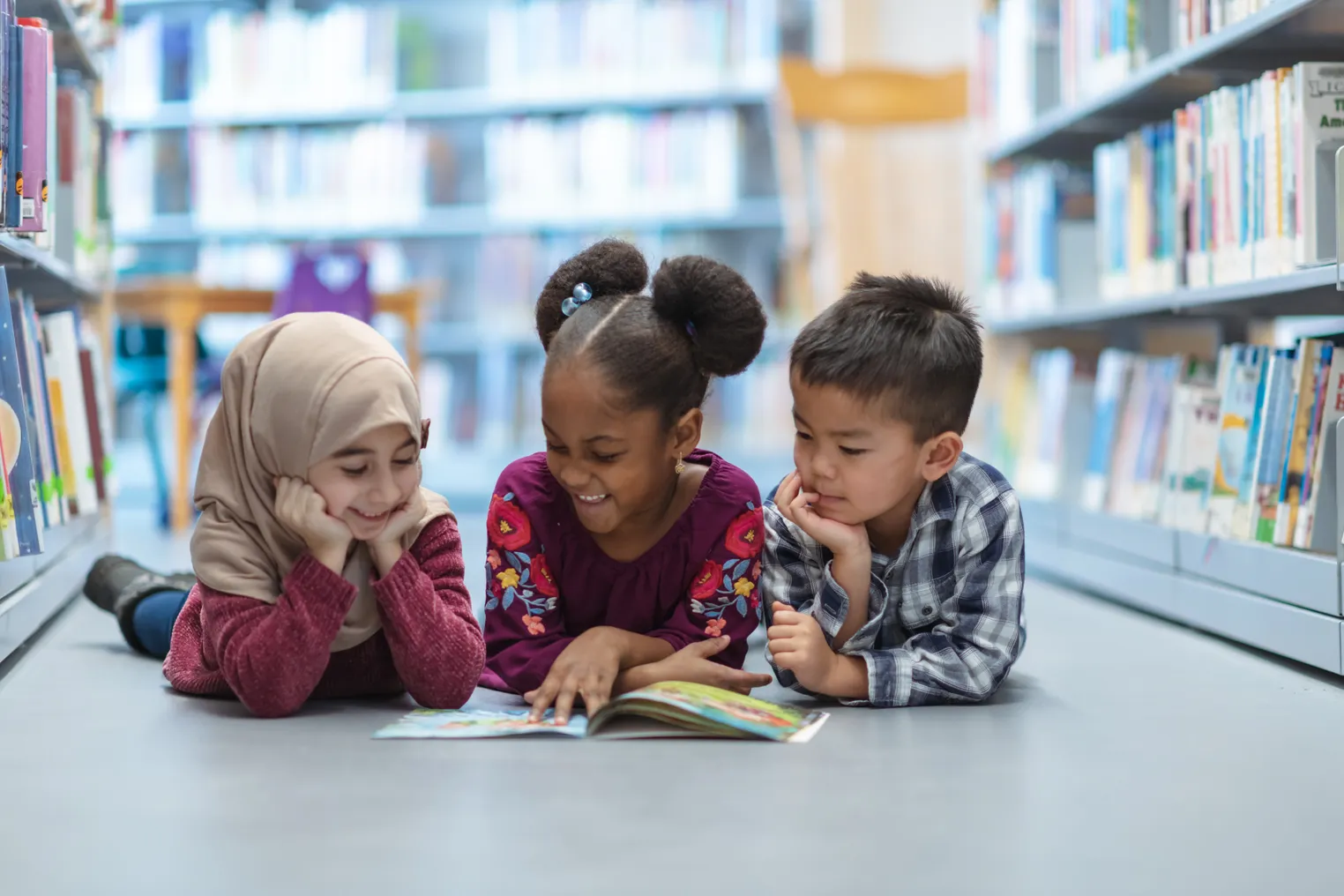 Three young children lie on their stomachs on the floor looking at an open book. There are rows of books on their side of them.