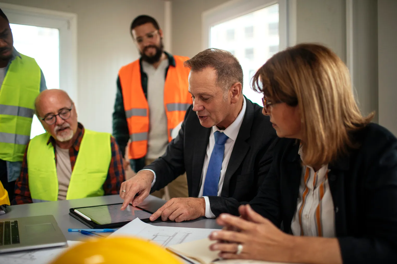 A group of construction stakeholders, some in suits, some in work gear, gather in a meeting.